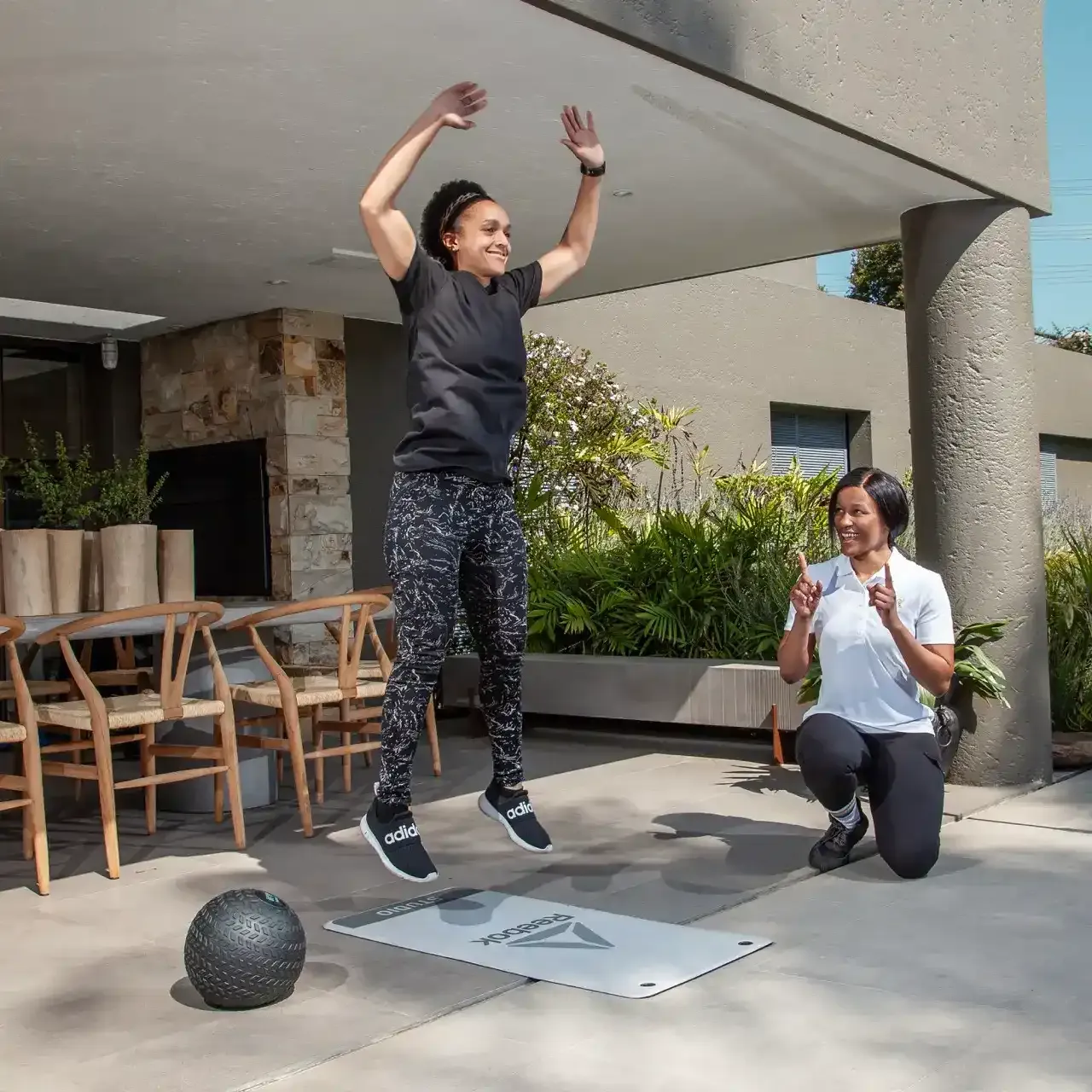Client leaping into the air during a jump squat while her trainer kneels and cheers her on