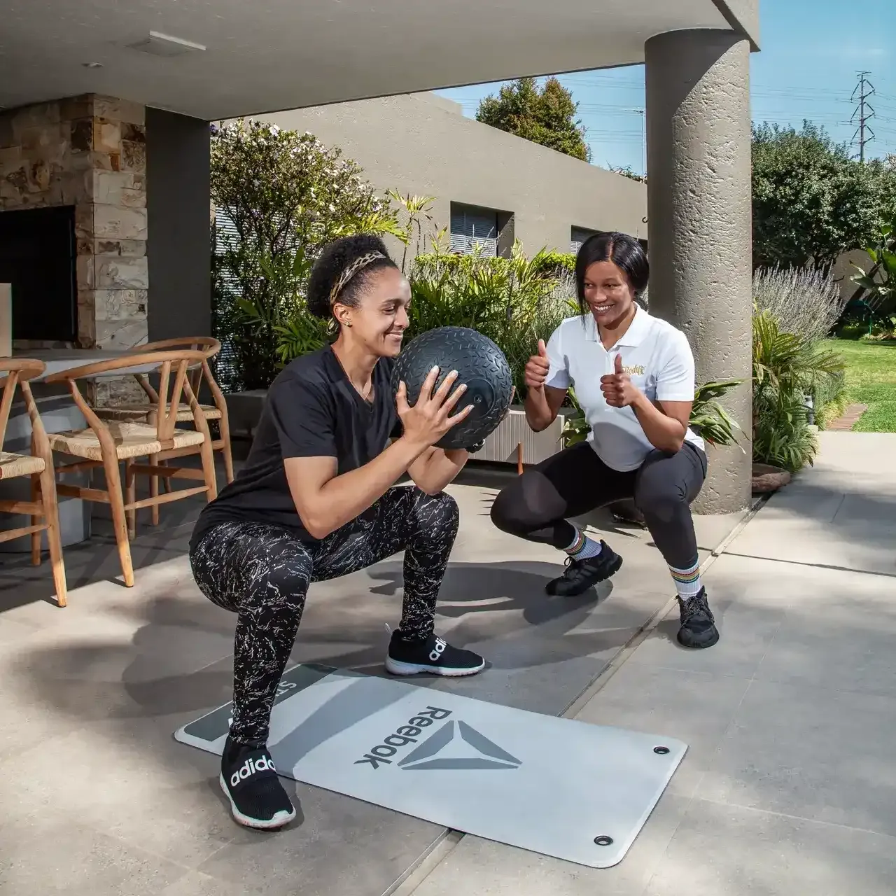 Client in a deep squat holding a slam ball as her trainer squats alongside giving two thumbs up