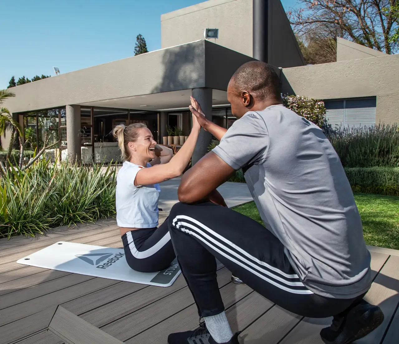 Client high fiving her trainer from an exercise mat on a wooden deck outside a modern home