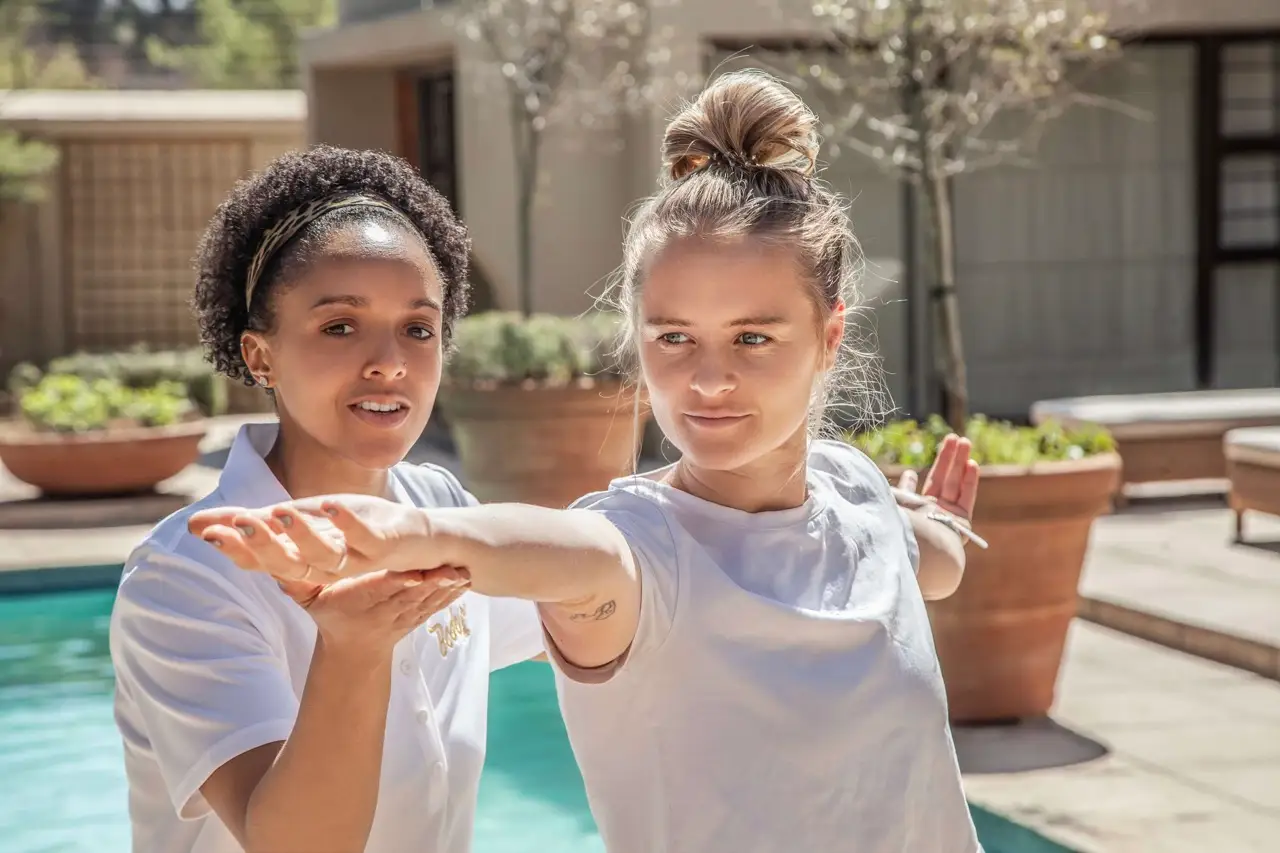 Trainer adjusting a client's outstretched arm in a warrior pose beside a garden pool