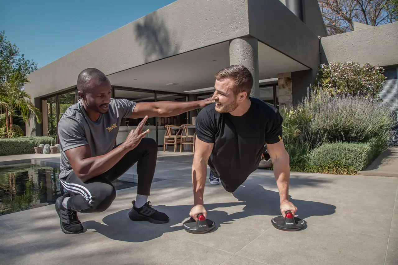 Trainer crouching beside a client doing push ups on rotating handles on a patio at a home