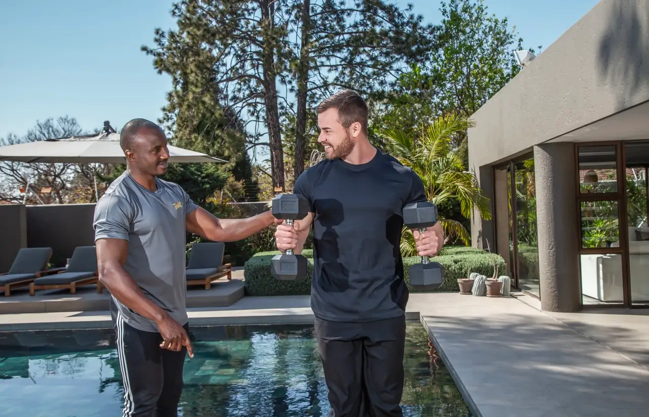 Trainer spotting a client holding two dumbbells beside a swimming pool at a modern home